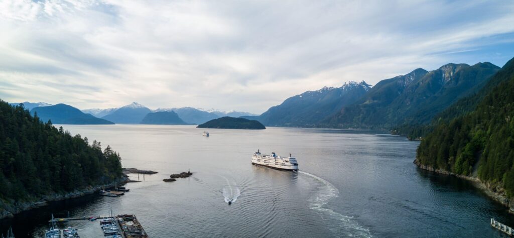 large ferry on water surrounded by mountains, sloped trees and billowy clouds.